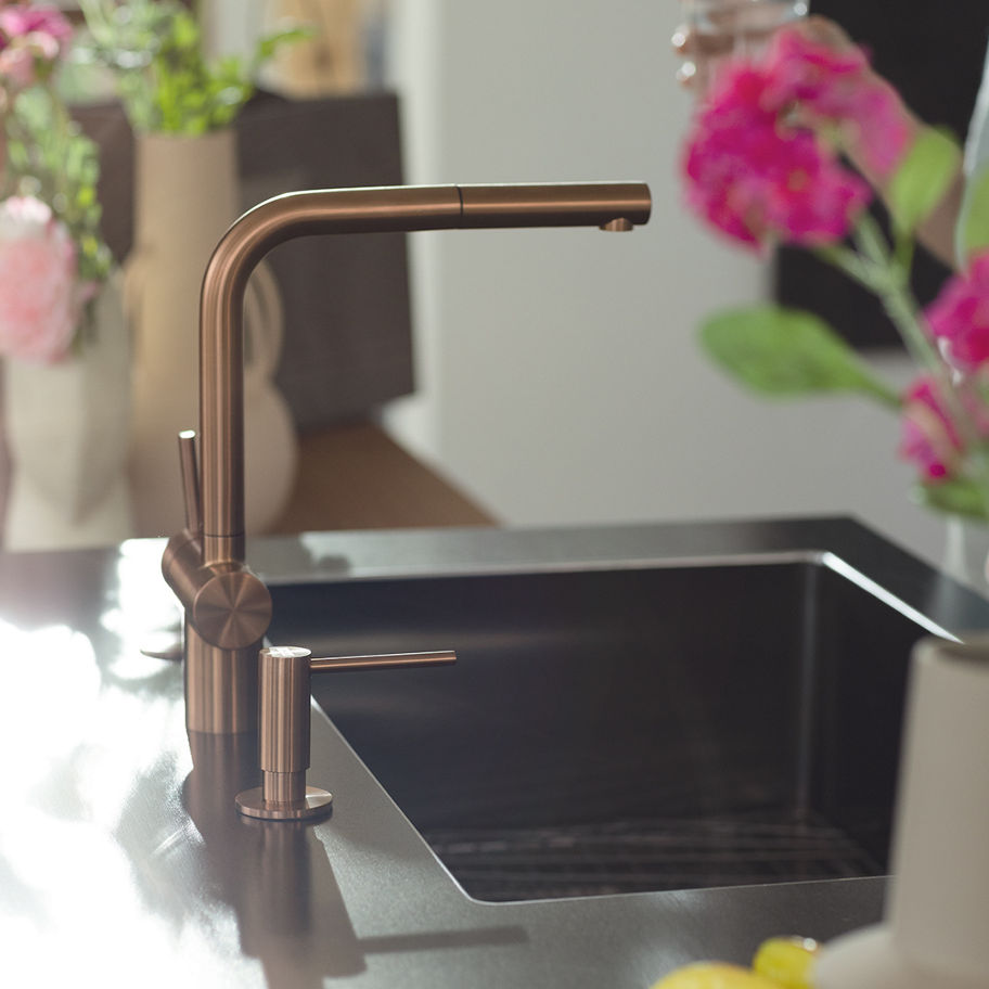 Close-up of a modern Franke kitchen faucet in a sleek, metallic finish, with a blurred background featuring pink and white flowers in vases and a dark countertop
